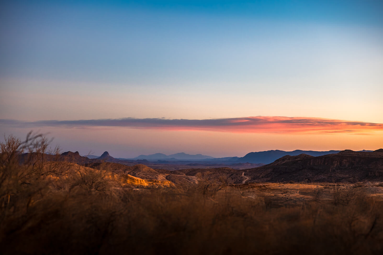 Terlingua Ghost Town Sunset — Big Bend Region, Texas Sweeping desert landscape at dusk near Terlingua Ghost Town, Texas. Golden scrub brush fills the foreground, layered mountain ridges fade into blue haze in the distance, and a dramatic band of pink and orange clouds stretches across a gradient sky — deep amber at the horizon bleeding into soft blue overhead.